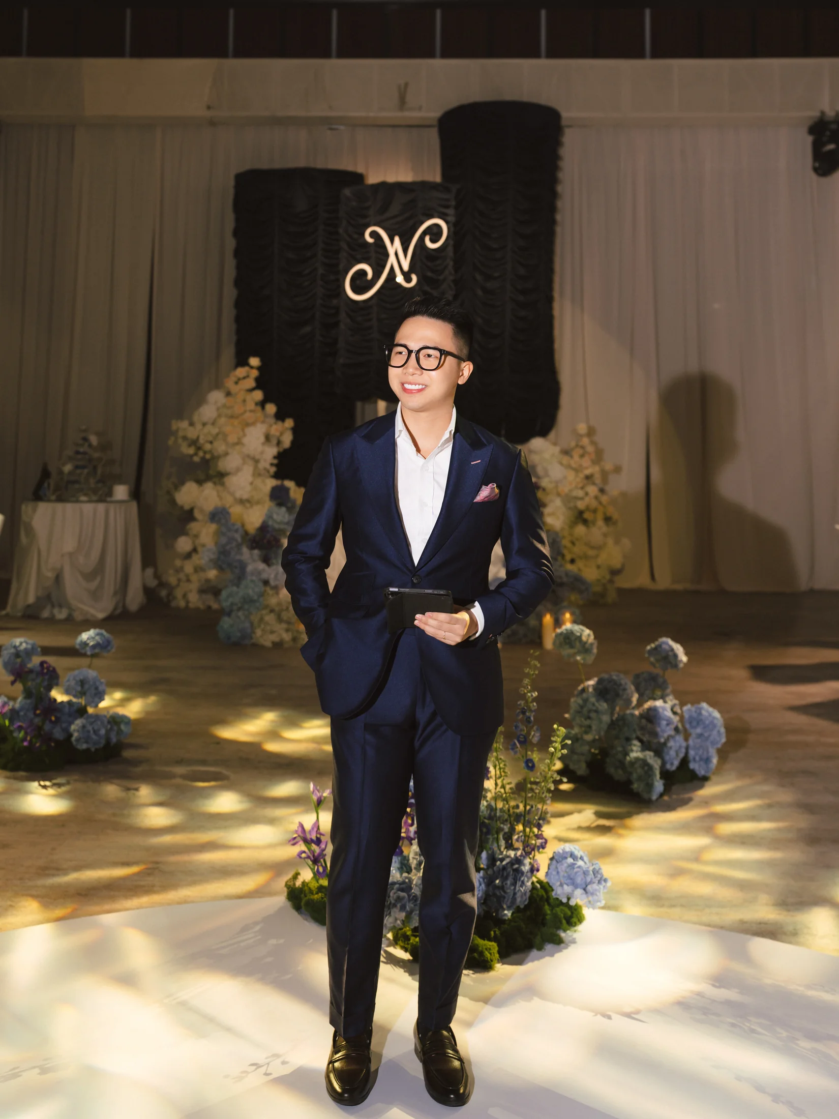 Chân dung MC Sơn Tùng mặc suit navy, mỉm cười và cầm kịch bản trong không gian lễ cưới